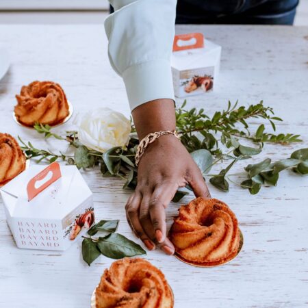 a hand grabbing a individual rum cake.on the table you also have 3 other rum cakes and 2 small packaging