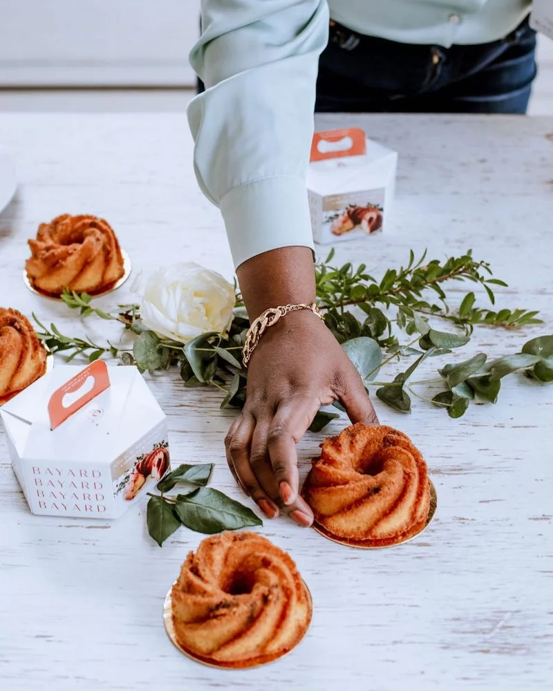 a hand grabbing a individual rum cake.on the table you also have 3 other rum cakes and 2 small packaging